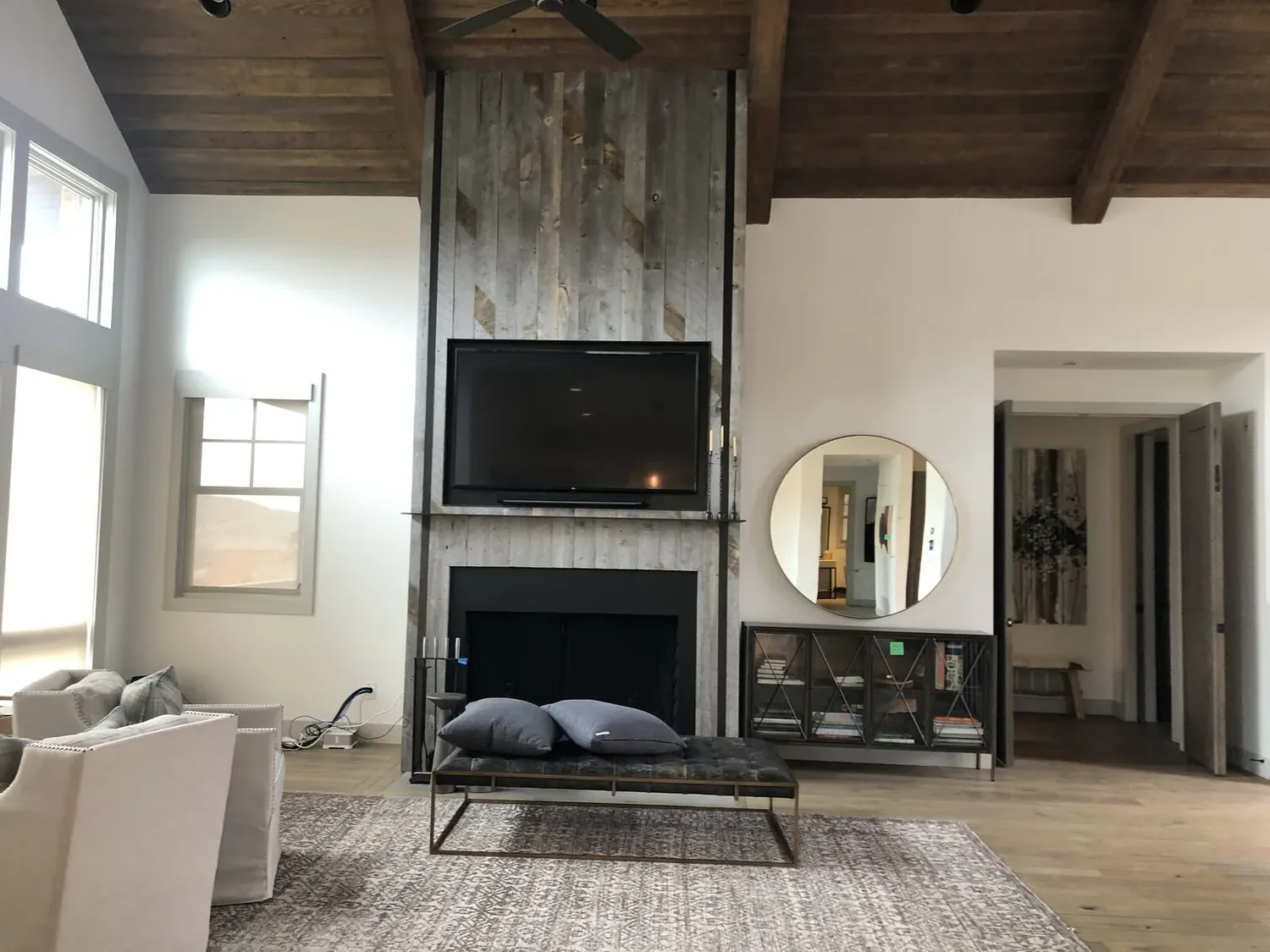 A modern living room with a mounted TV above a rustic wood-paneled fireplace, a bench with pillows, a round mirror