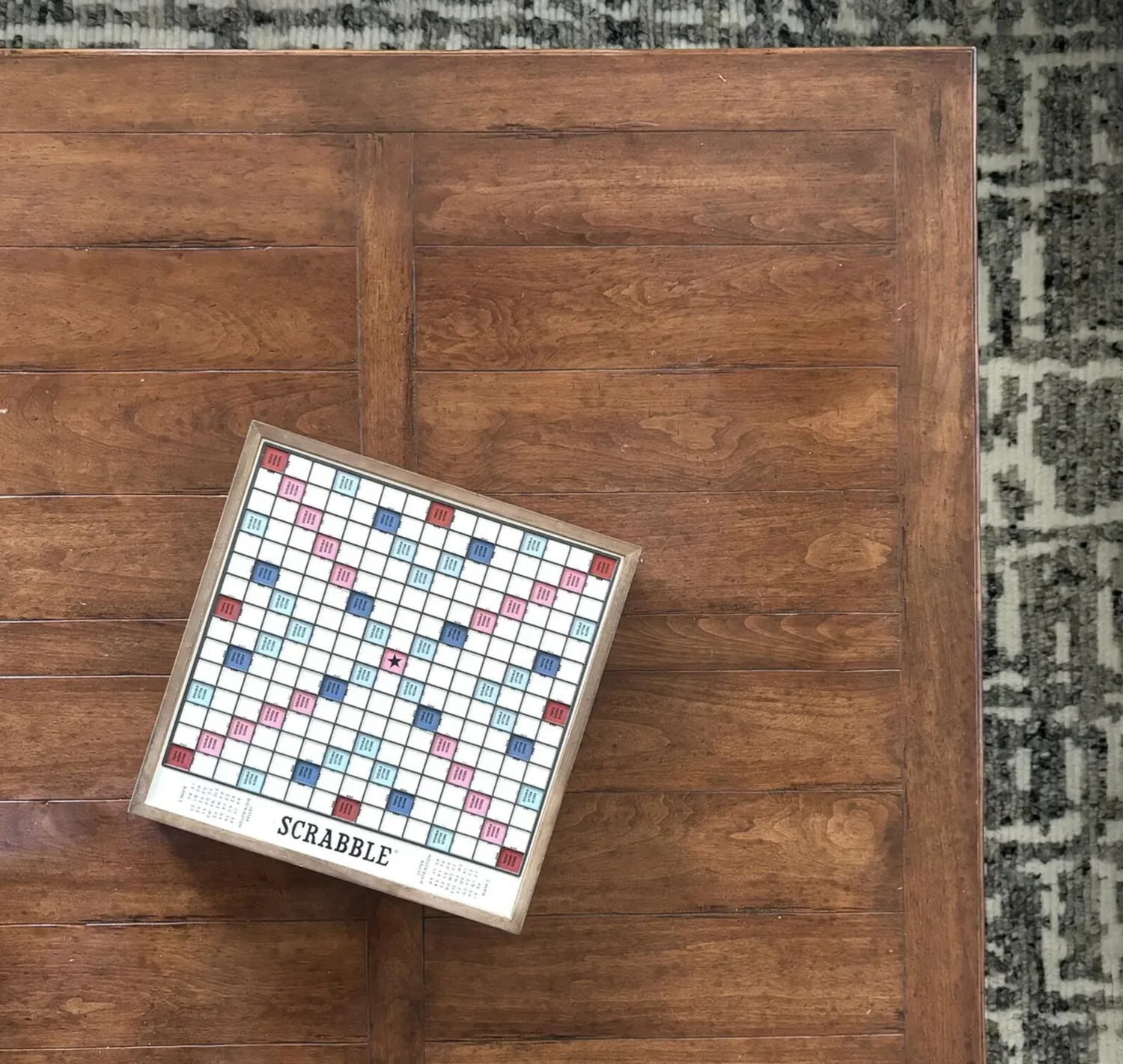 A wooden coffee table with a Scrabble board placed on top, next to a patterned rug