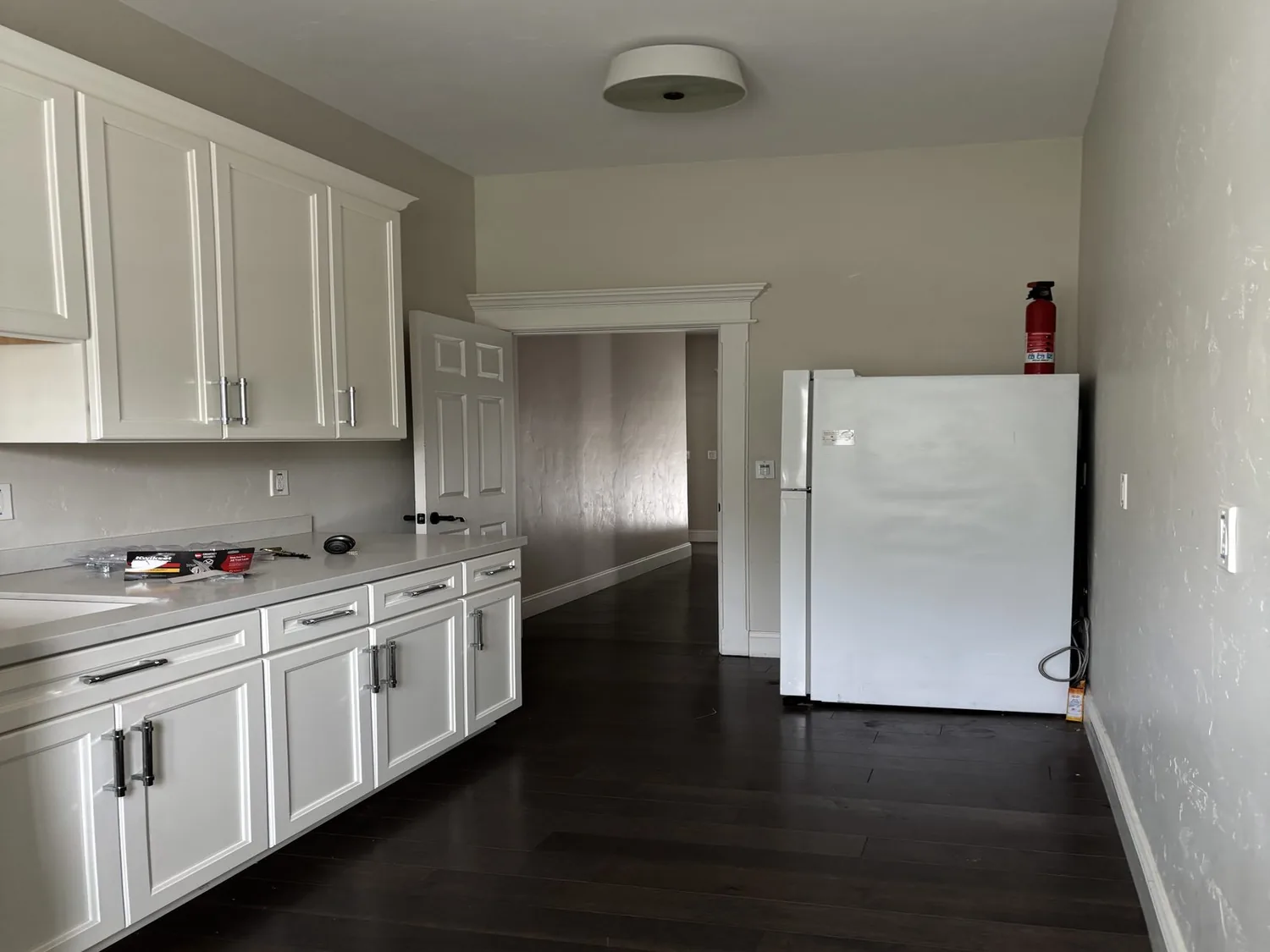 A kitchen with white cabinets, a white refrigerator with a fire extinguisher on top, dark wood flooring, and a beige