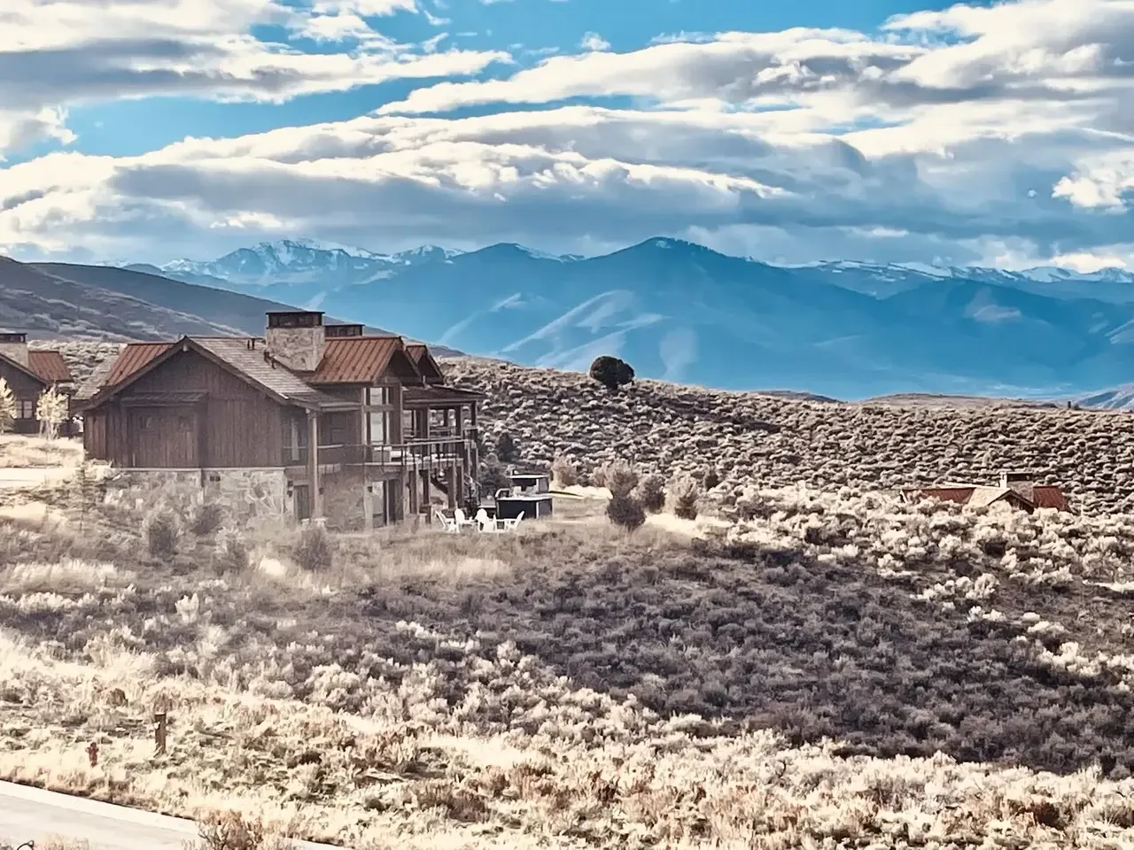 A rustic house with stone and wood exterior situated in a dry, shrub-covered landscape with mountains in the background
