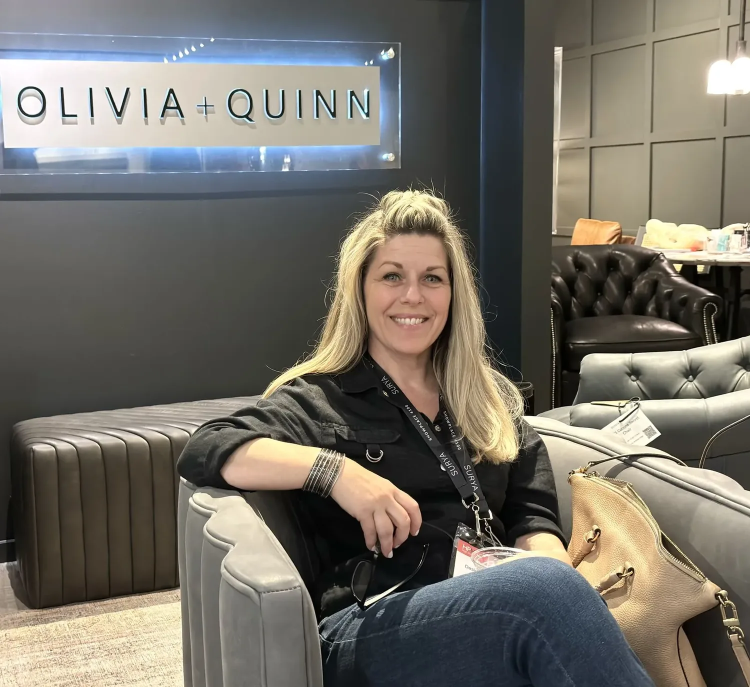 A woman sitting on a gray tufted sofa in a modern showroom with a sign reading OLIVIA + QUINN in the background
