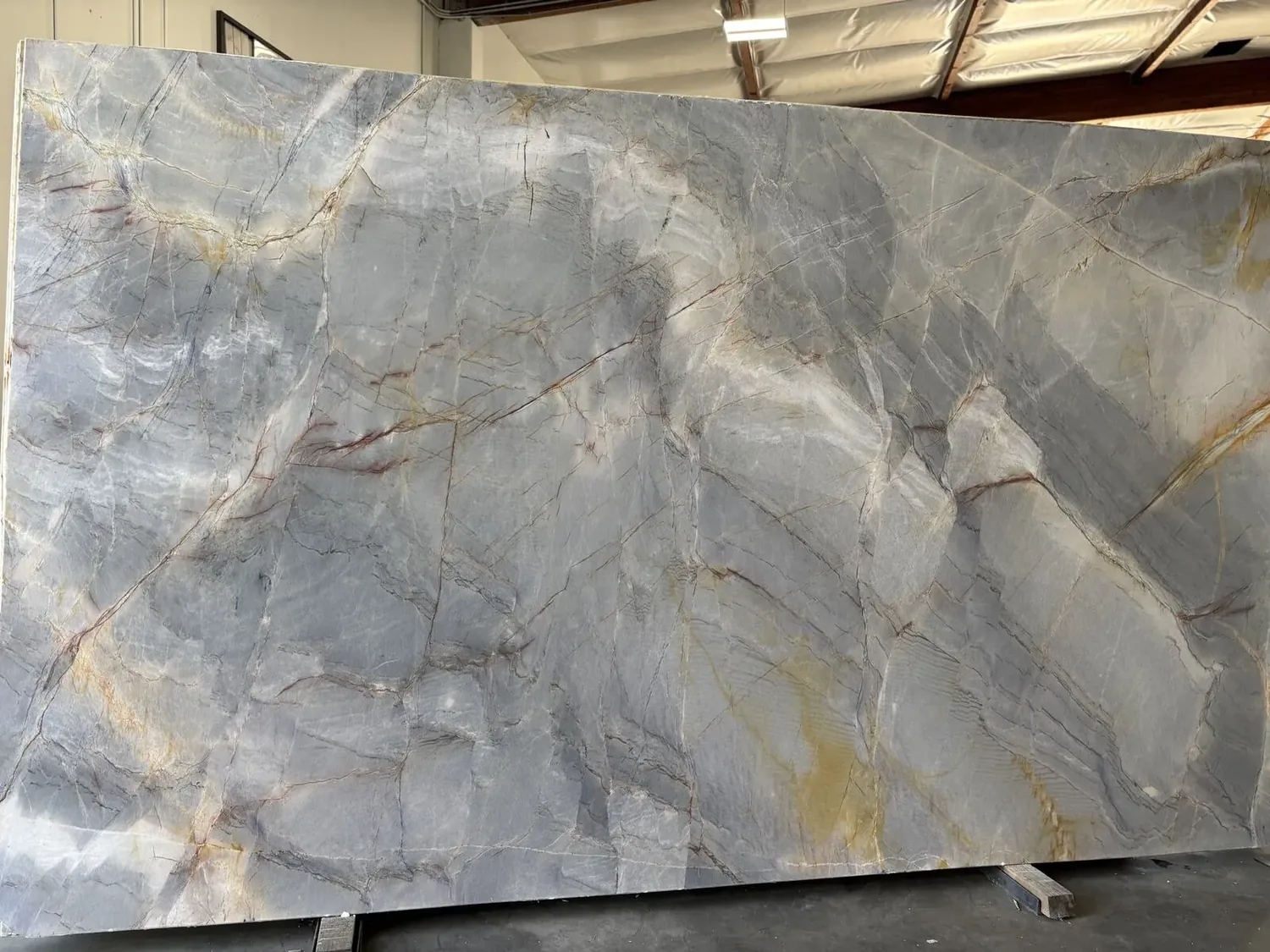 A large slab of natural stone with gray, white, and yellowish veining, displayed indoors on a stand