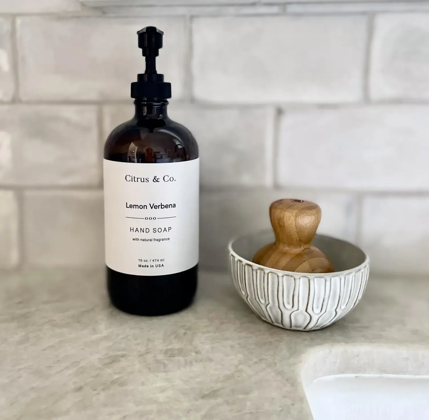 A bottle of Citrus & Co. Lemon Verbena hand soap and a wooden soap holder in a decorative bowl on a bathroom countertop