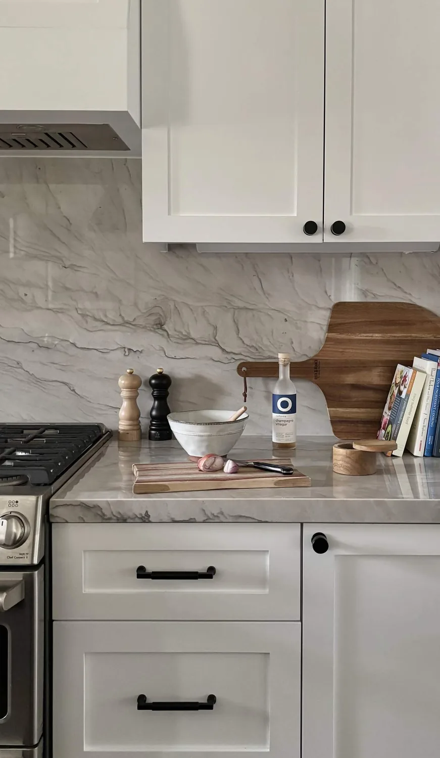 A modern kitchen countertop with white cabinets, marble backsplash, a cutting board, a bowl, pepper mills, a bottle of champagne vinegar, and cookbooks