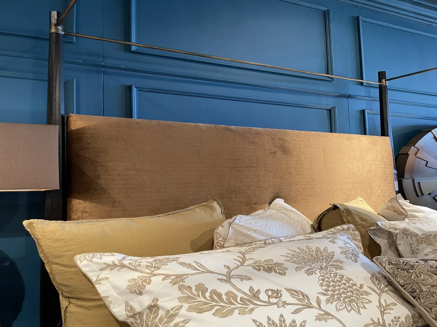 A close-up of a bed with a brown upholstered headboard, beige and patterned pillows, set against a blue paneled wall