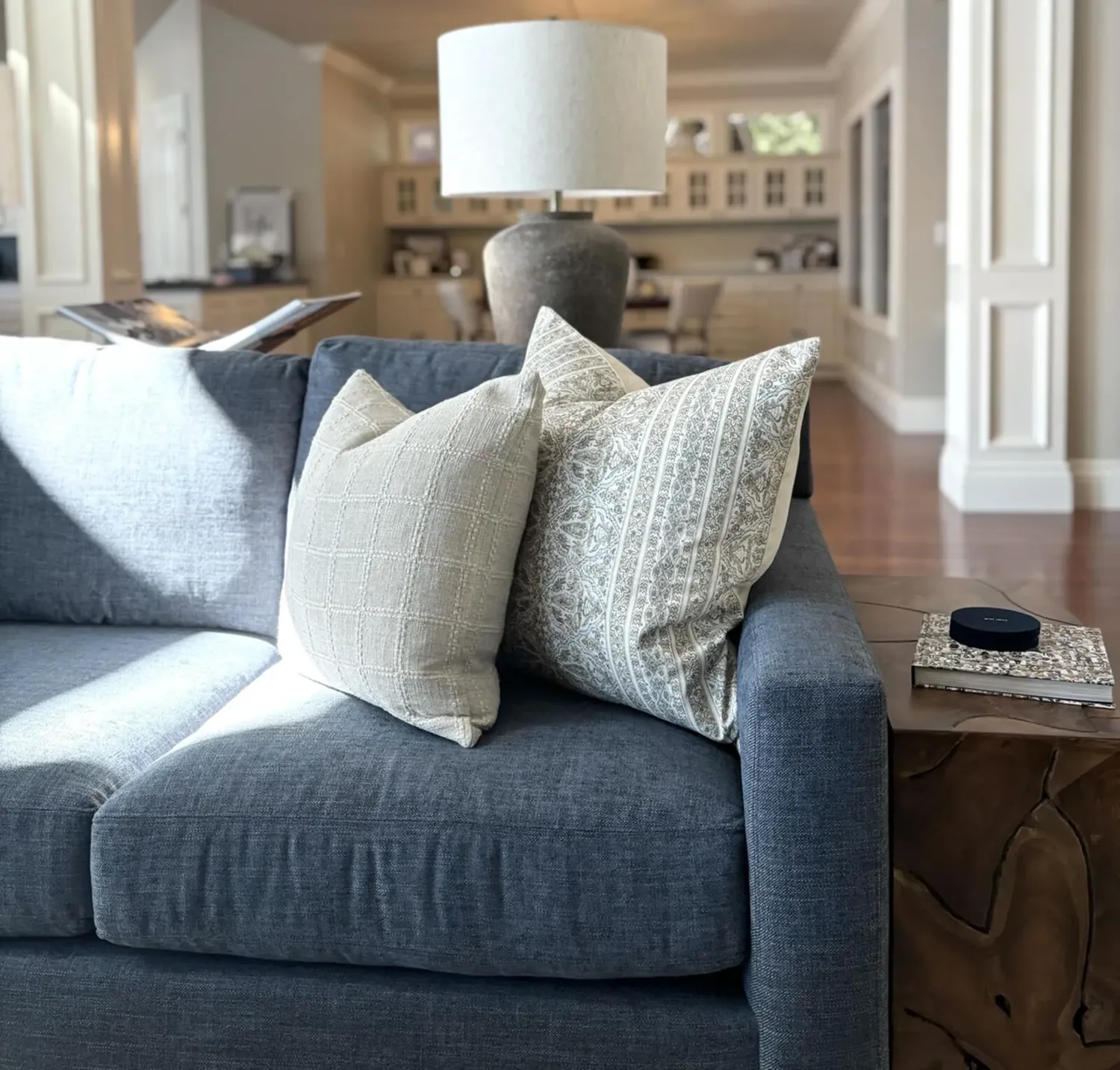 A modern living room with a blue sofa adorned with patterned pillows, a large lamp, and a wooden side table with books