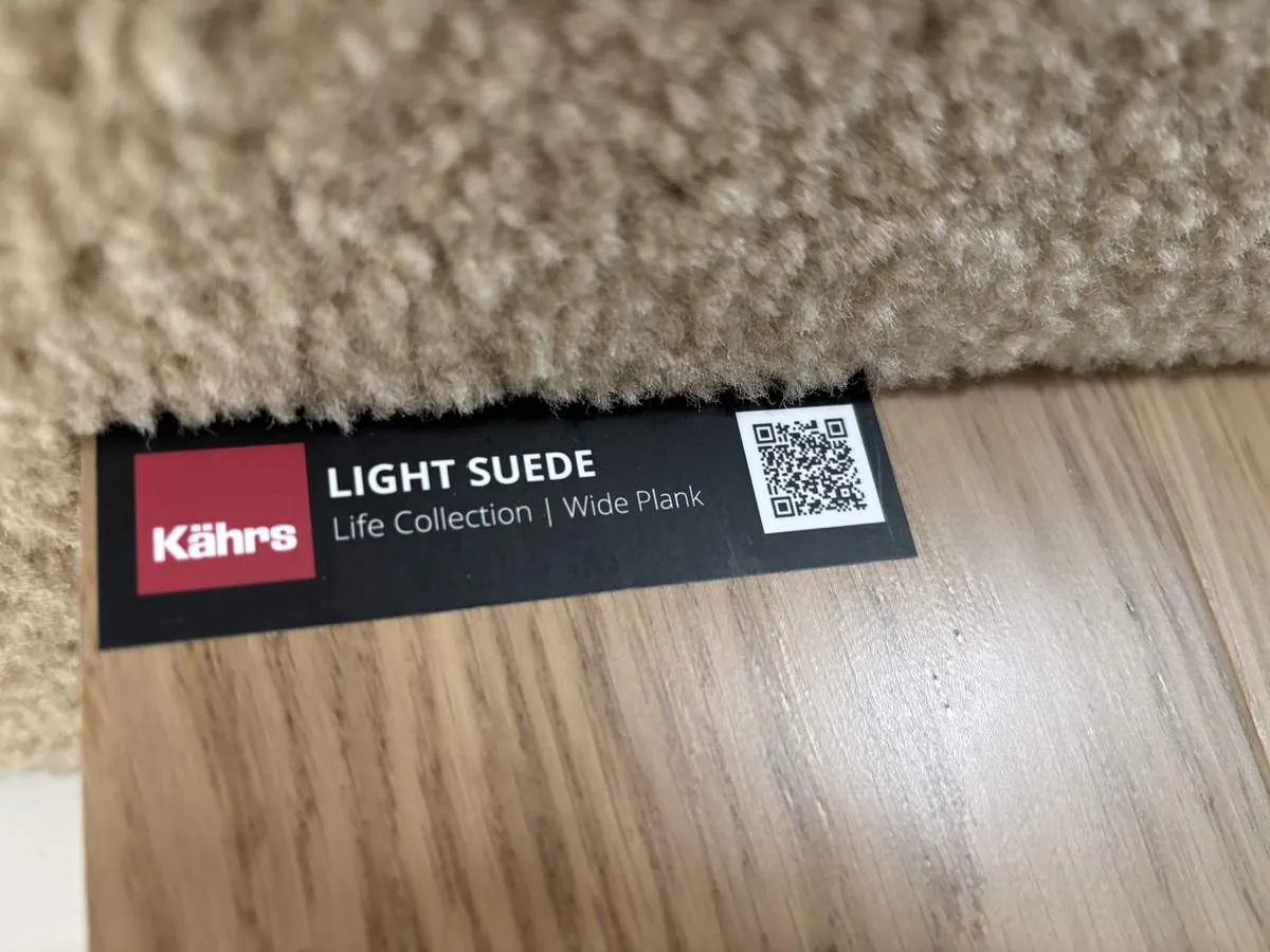 Close-up of a flooring sample labeled 'Light Suede' from Kährs Life Collection with a beige carpet edge