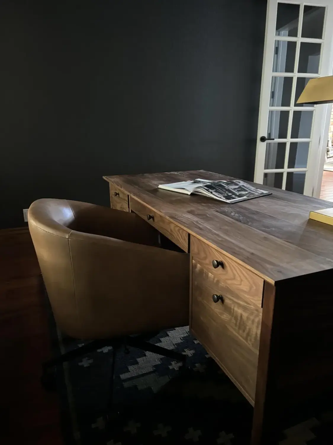 A wooden desk with drawers and a brown leather chair in a dark room with a window and patterned rug