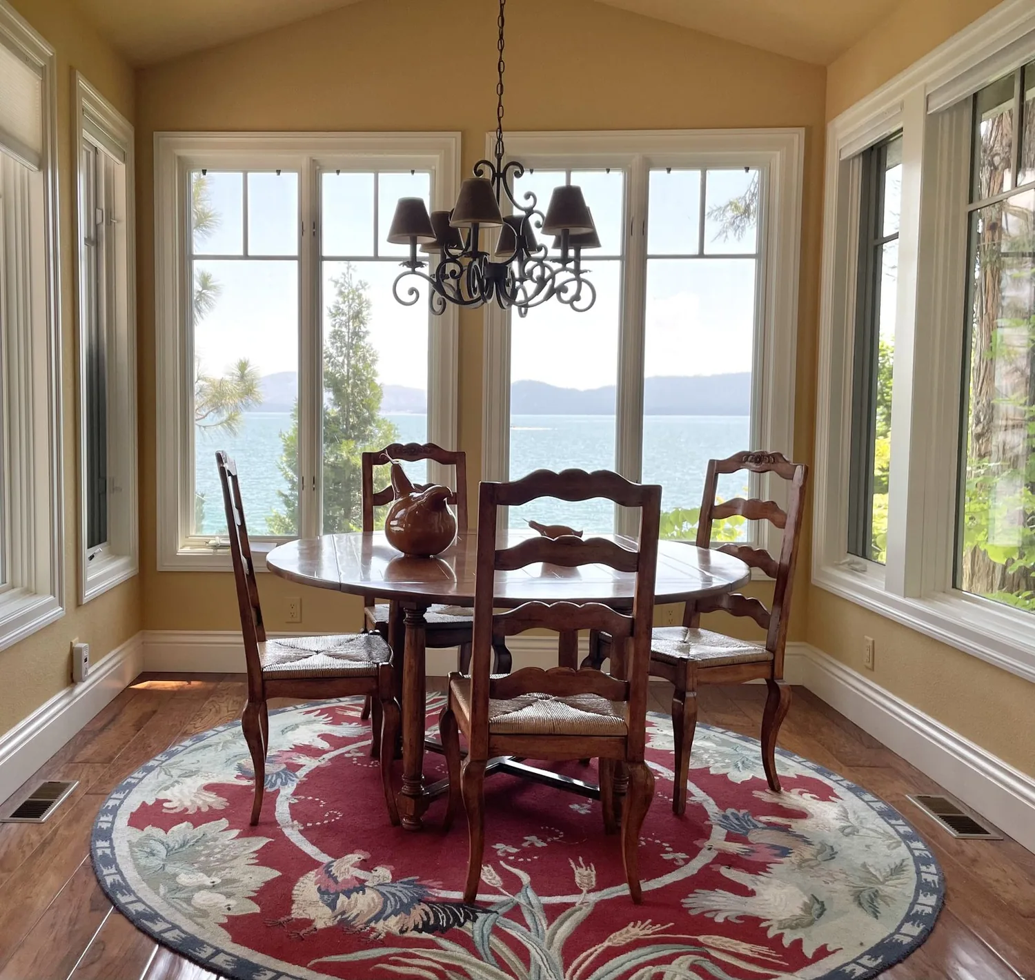 A dining room with a round wooden table, four wooden chairs, a decorative chandelier, and large