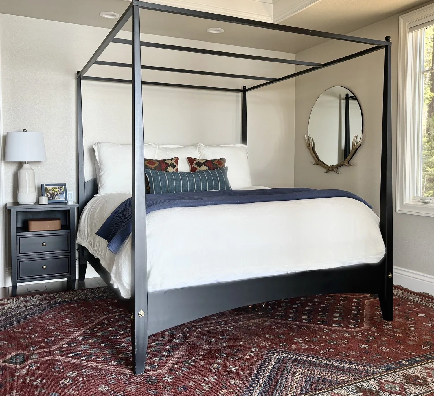 A bedroom with a black metal canopy bed, white bedding, patterned pillows, a nightstand with a lamp and photo frame