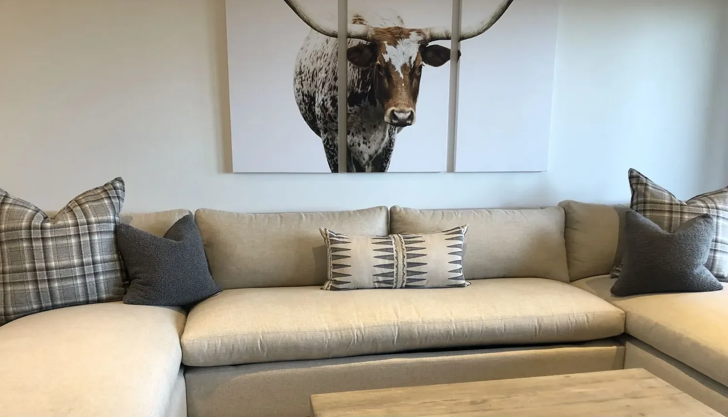 A beige sectional sofa with patterned and solid pillows beneath a three-panel wall art of a longhorn steer in a living