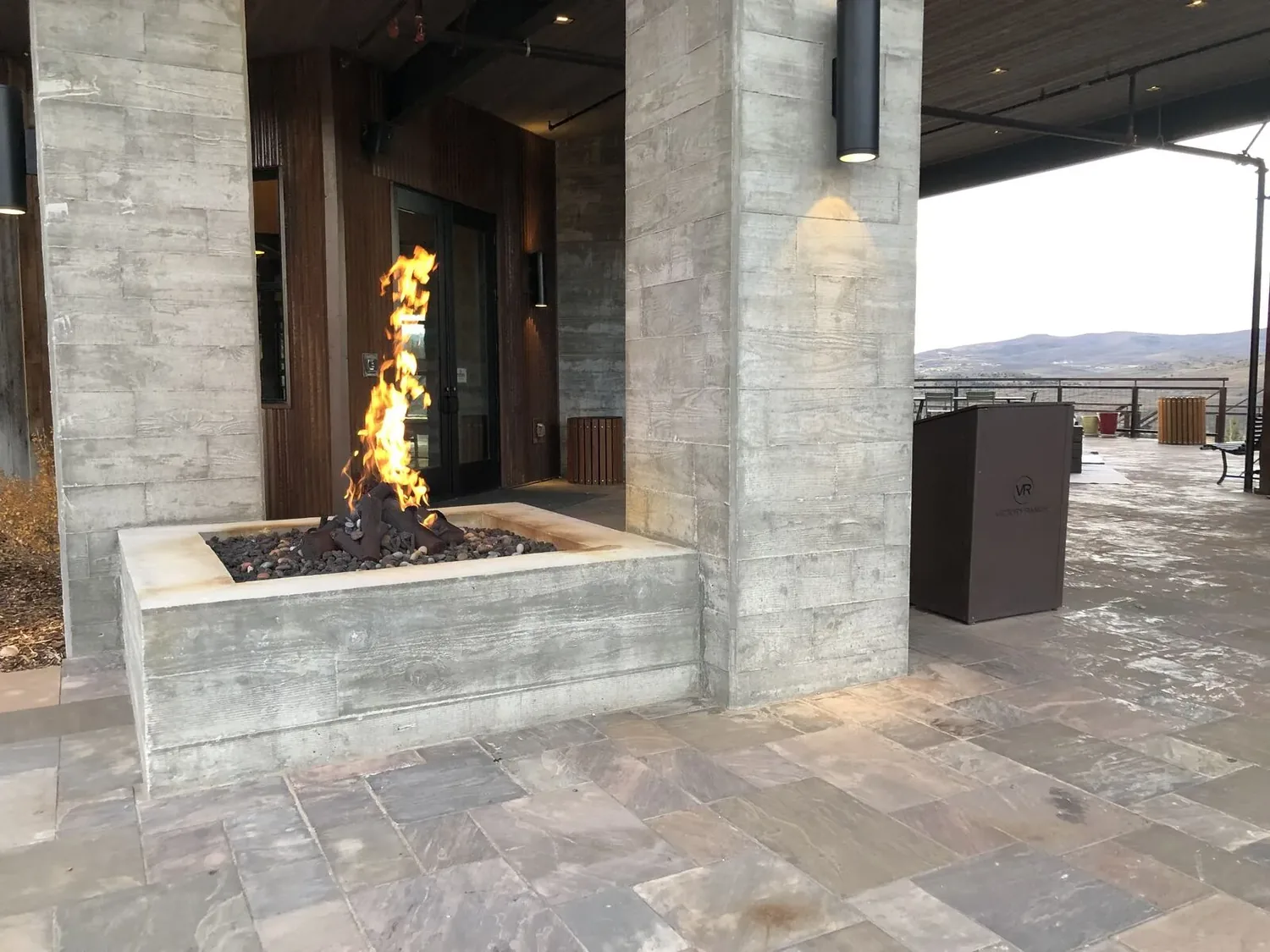 Outdoor patio area with a modern concrete fire pit and stone tile flooring, overlooking a mountainous landscape