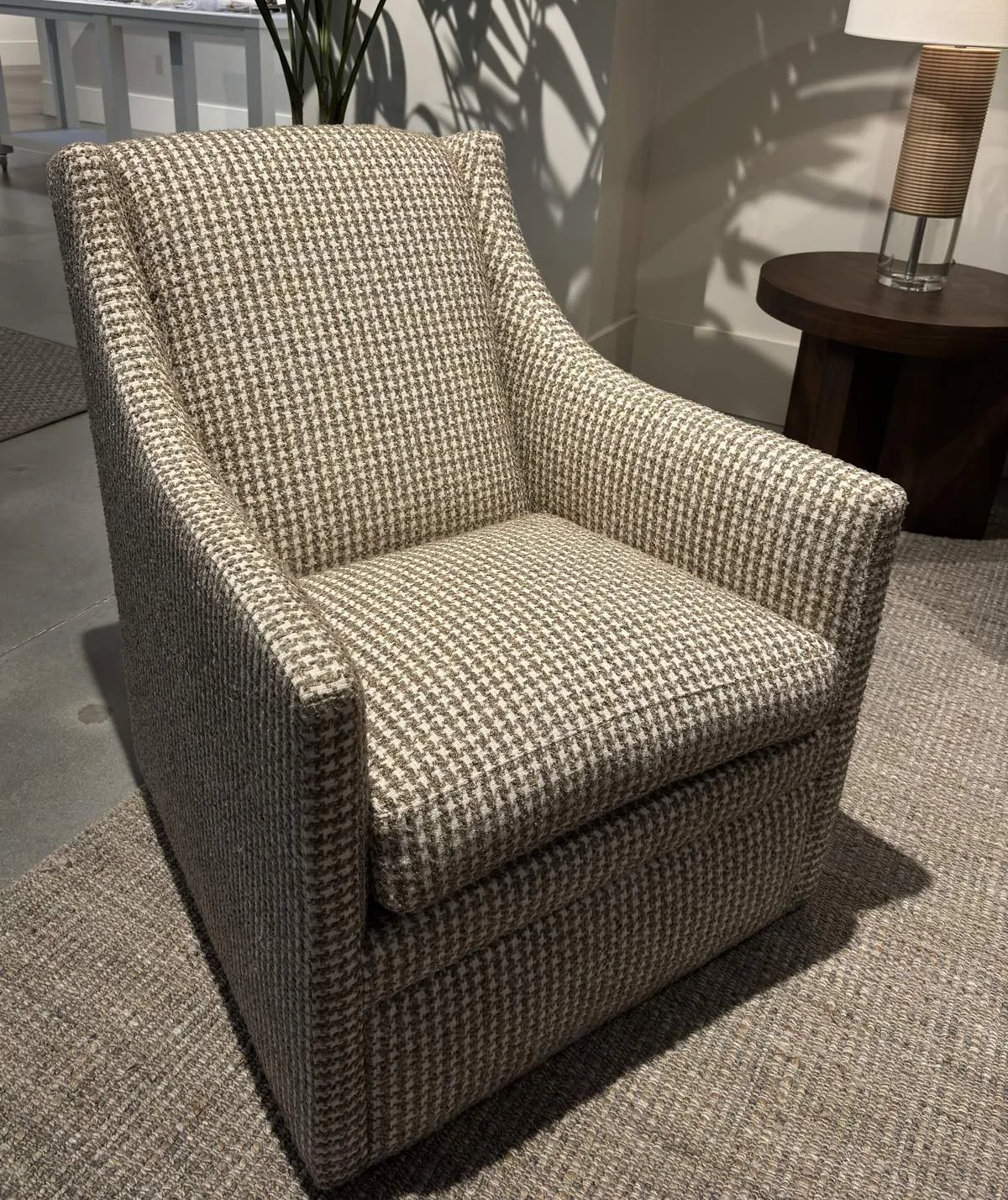 A beige and brown houndstooth patterned armchair placed on a textured rug with a wooden side table and lamp in the background