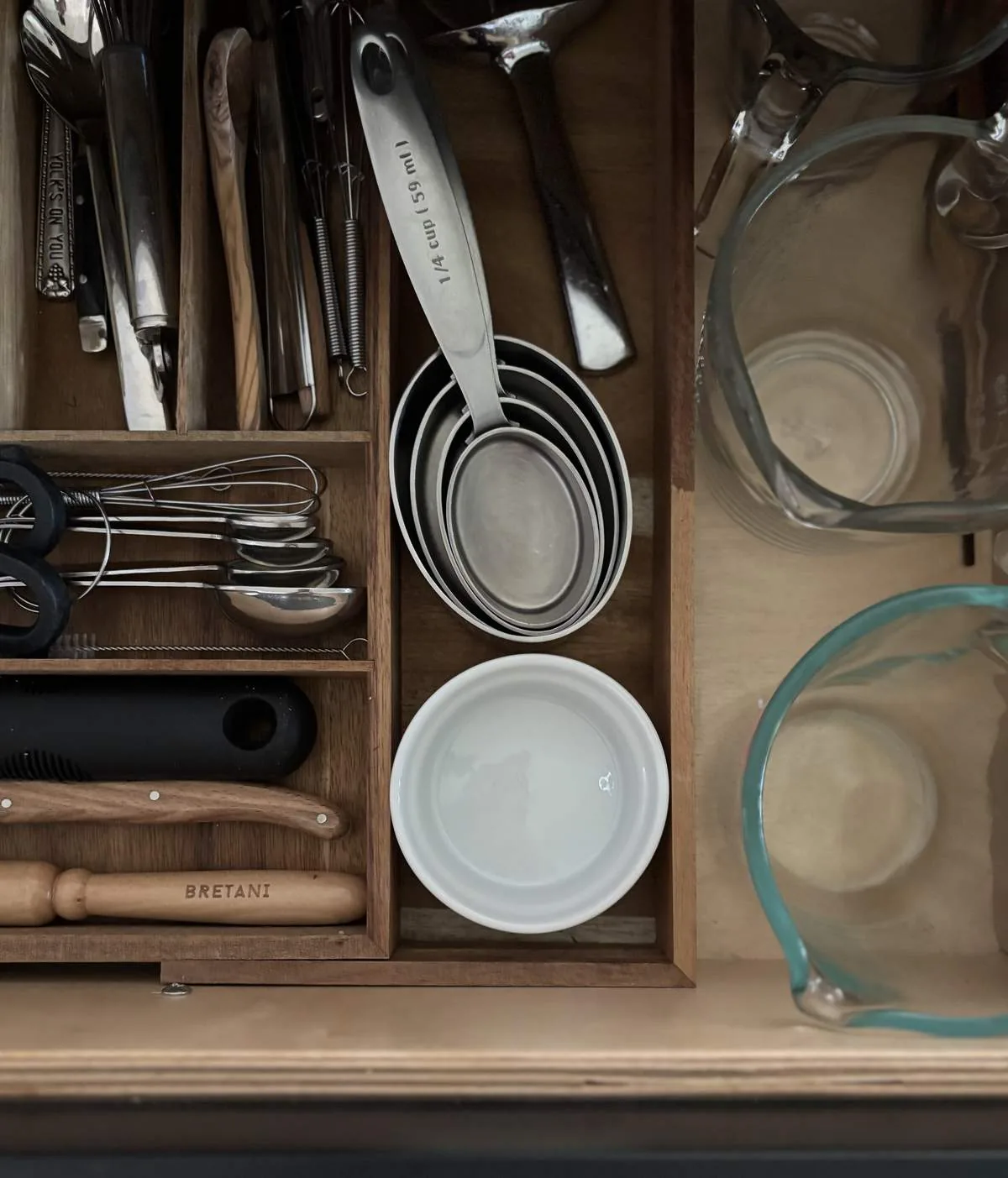 Top-down view of a wooden kitchen drawer organizer containing measuring spoons, whisks, knives, and glass measuring cups