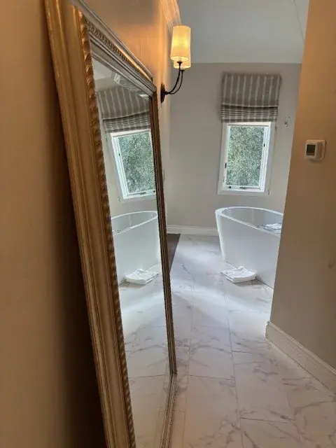 A bathroom with a freestanding bathtub, marble floor tiles, a large framed mirror, and a window with striped curtains