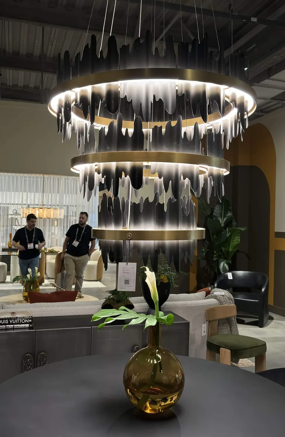 A modern interior space featuring a large, artistic chandelier with black and white dripping design, a round table with a brown glass vase holding a white flower, and contemporary furniture in the background
