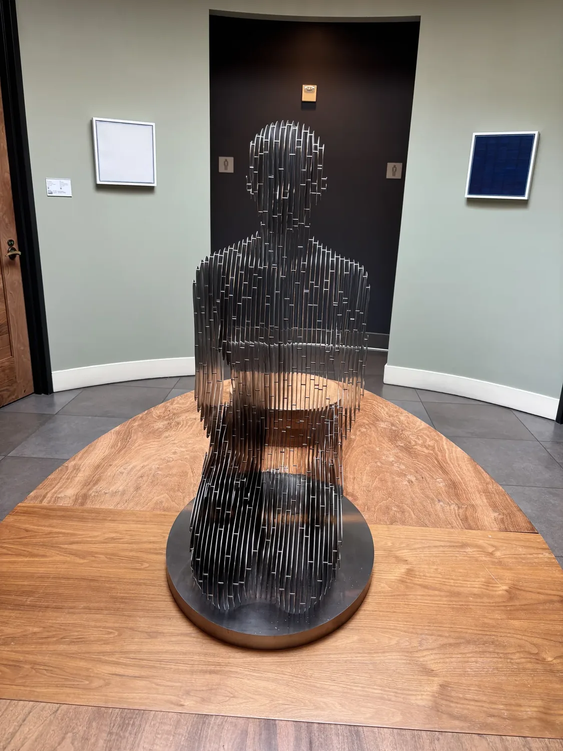 A modern art sculpture made of vertical metal rods forming a human figure, displayed on a wooden table in a gallery space with light green walls and framed artworks