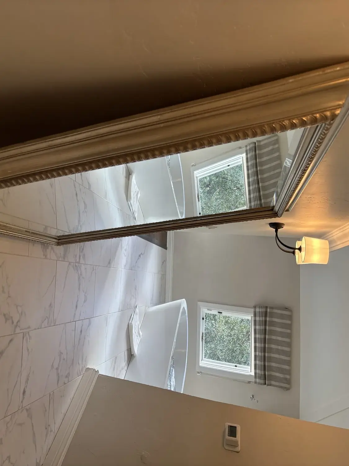 A modern bathroom with a freestanding bathtub, large mirror, wall sconce, and marble tile flooring