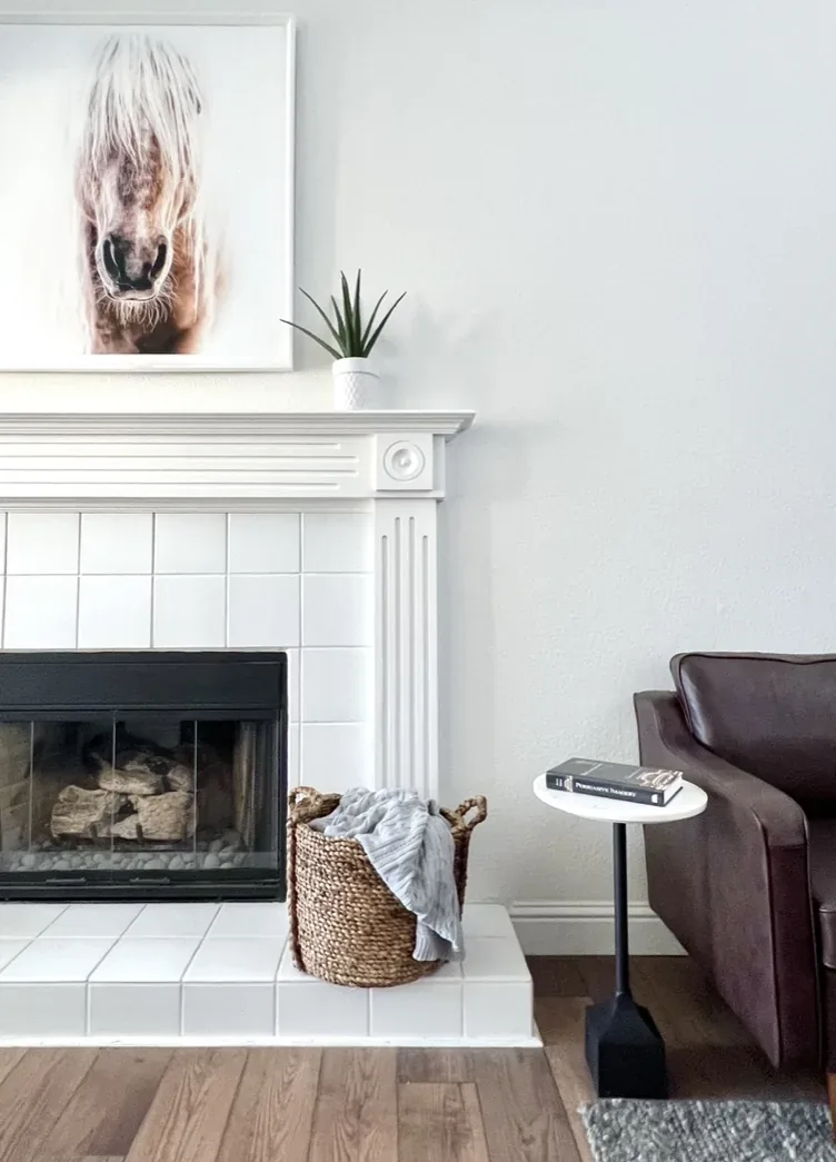 A modern living room corner with a white tiled fireplace, a woven basket with a blanket, a small side table with books, and a dark leather armchair