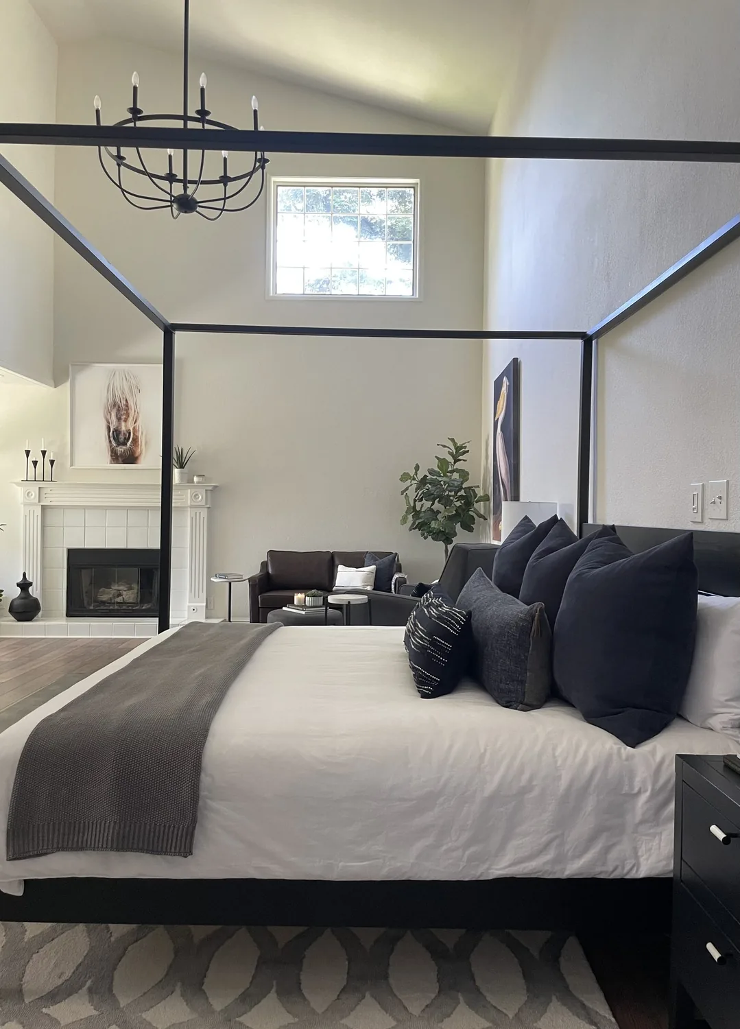 A modern bedroom with a black four-poster bed, dark pillows, a gray throw, a sitting area with a dark sofa