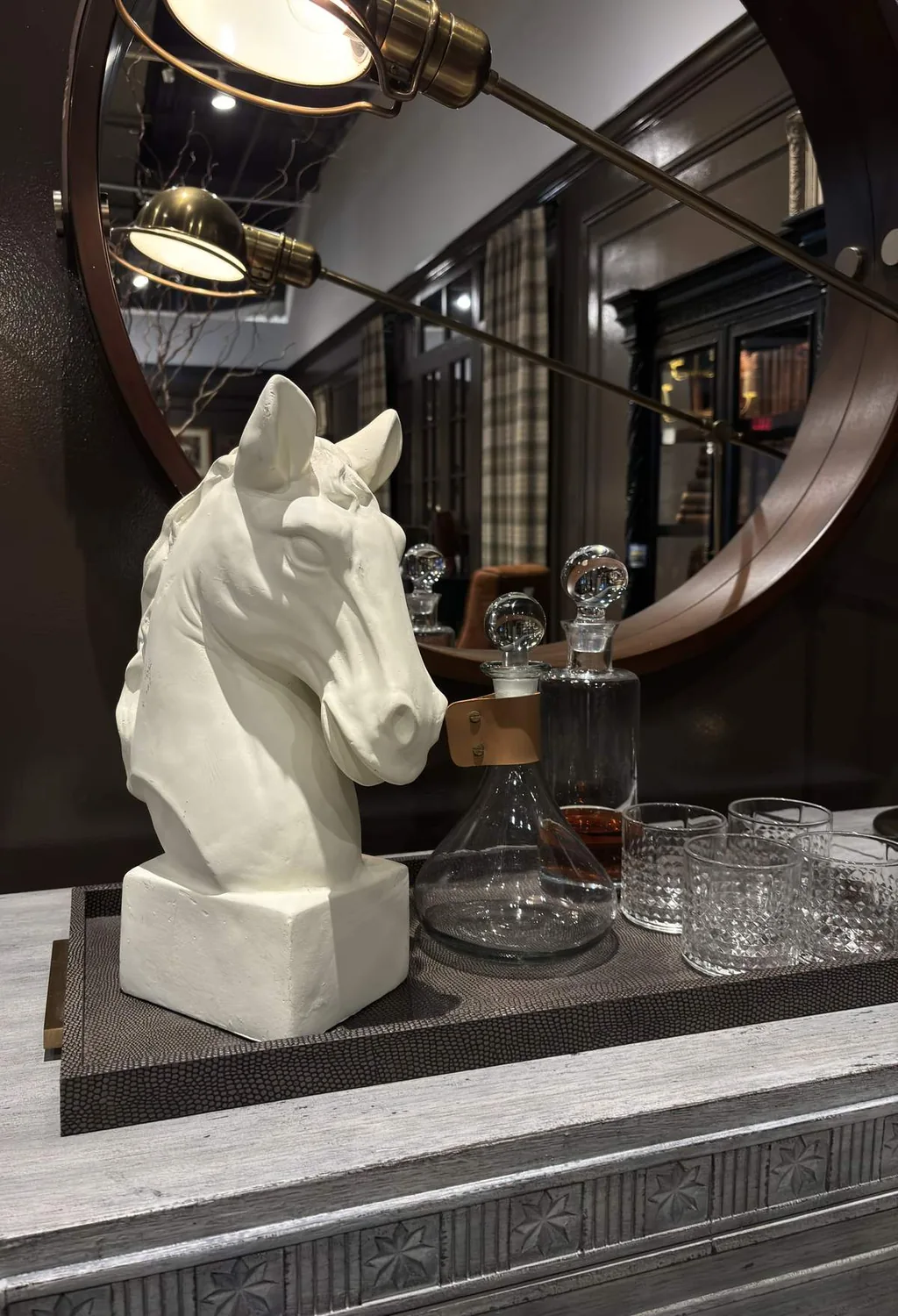 A decorative arrangement featuring a white horse head sculpture, glass decanters, and glasses on a tray in front of a round mirror with a brass lamp reflected
