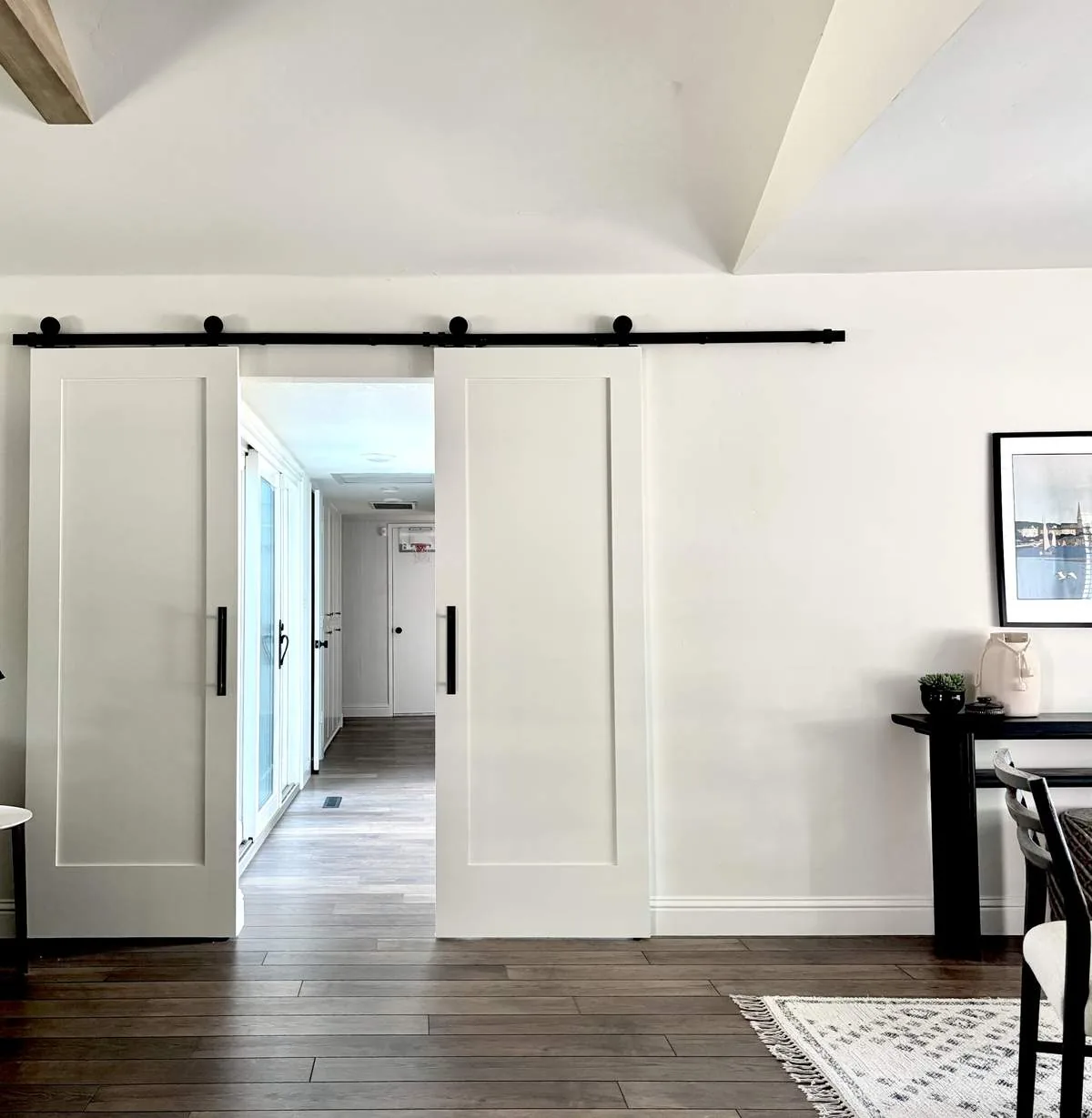 A modern interior space featuring white sliding barn doors on a black track, wood flooring, and a partial view of a dining area with a black table and framed artwork