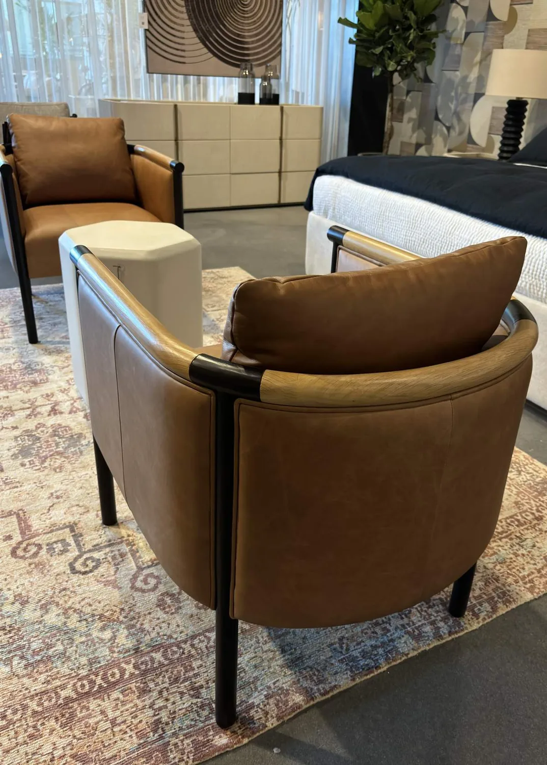 A modern bedroom seating area featuring two brown leather chairs, a white geometric side table, a patterned rug, and a bed with a dark blue blanket