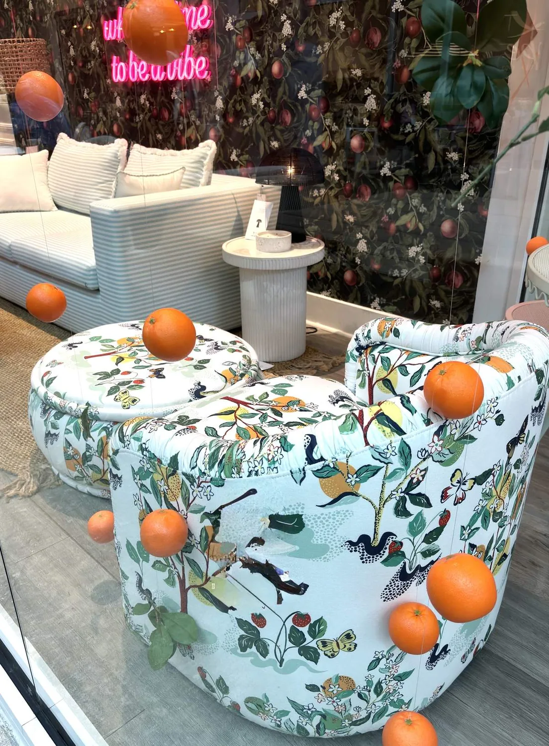 A cozy living room setup with floral patterned chairs, a striped sofa, a small round side table, and a dark floral wallpaper background, accented with floating oranges and a neon sign