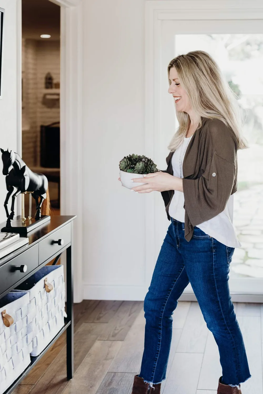 A woman holding a small potted succulent plant in a bright, modern living space with wooden flooring and a black console table with white baskets