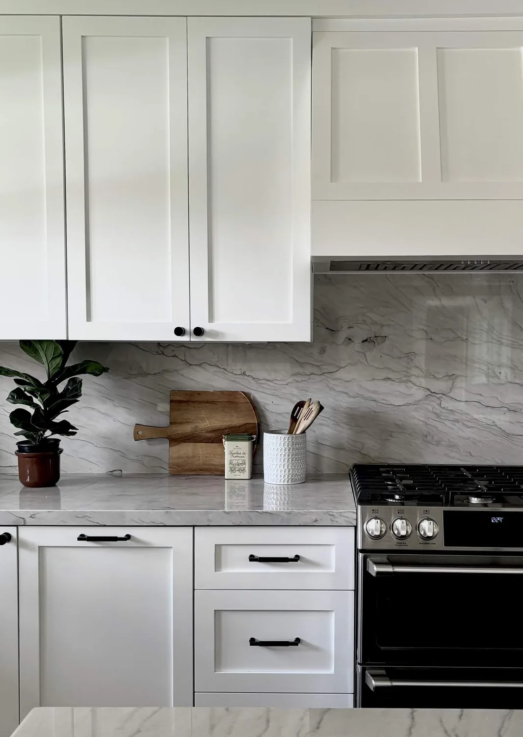 A modern kitchen with white cabinetry, marble countertops and backsplash, a stainless steel stove, and kitchen utensils on the counter