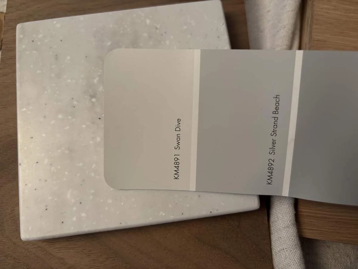 A paint color swatch card showing two shades of gray placed on a speckled white tile on a wooden surface