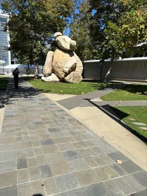 Outdoor scene featuring a large stone sculpture of a bear sitting on grass near a paved walkway with trees and buildings in the background
