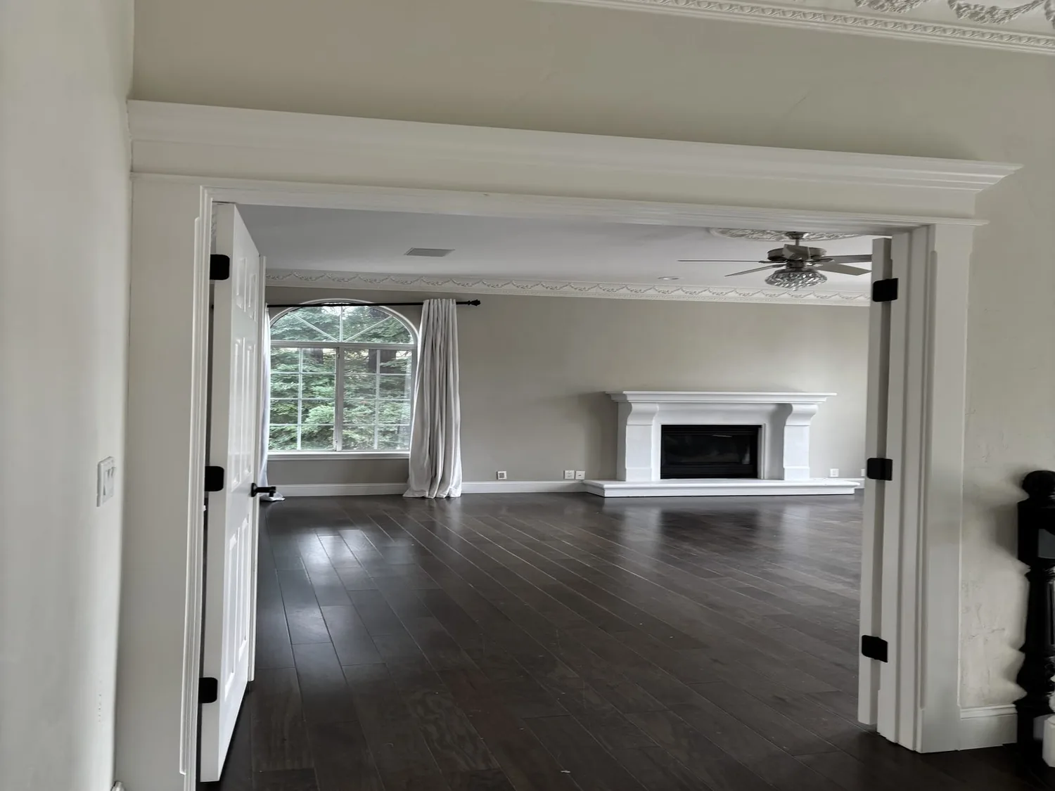 Empty living room with dark hardwood floors, a white fireplace, large arched window with curtains, and ceiling fan