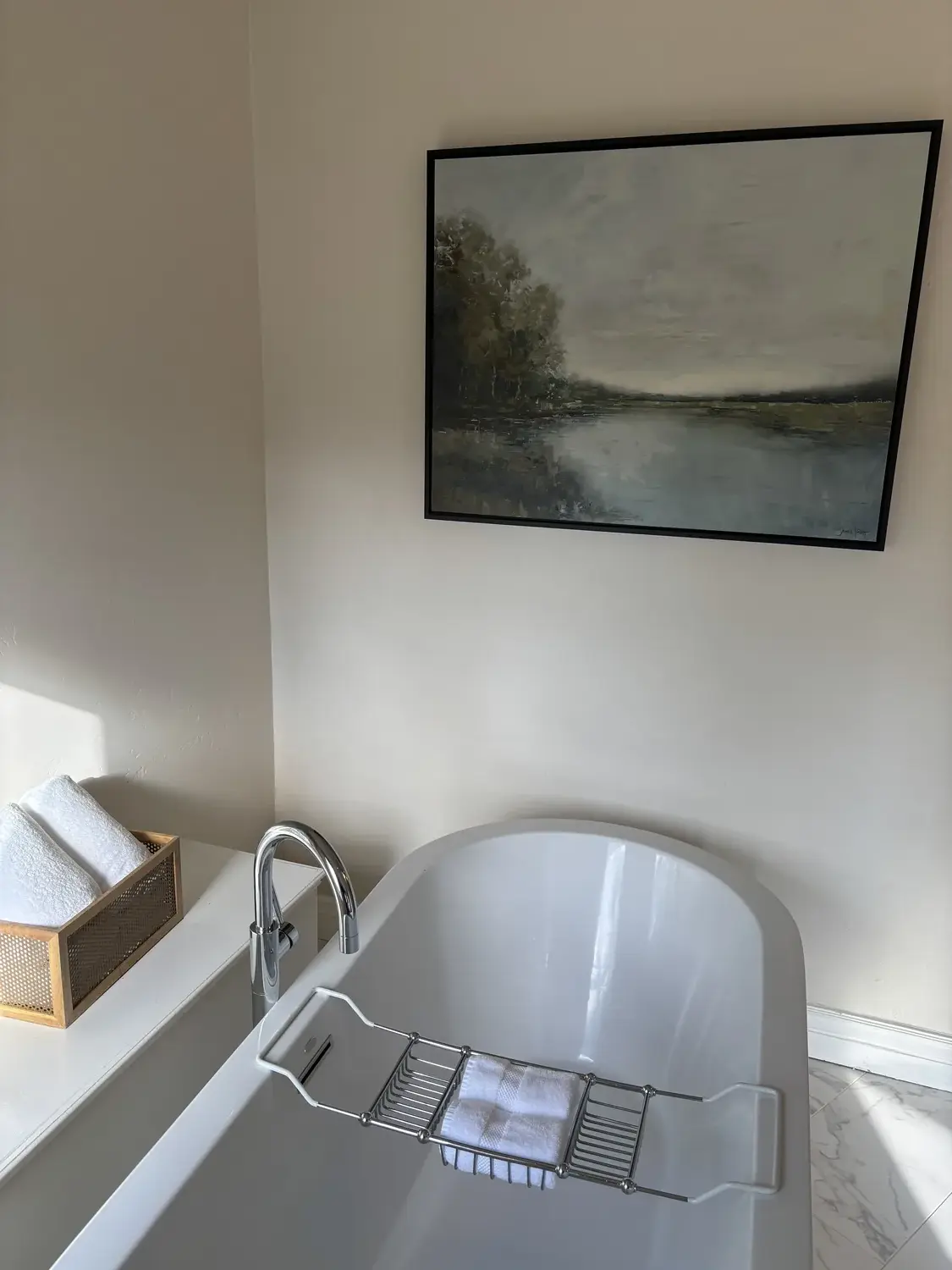 A modern bathroom featuring a white freestanding bathtub with a chrome faucet, a towel rack with white towels, and a large abstract painting on the wall