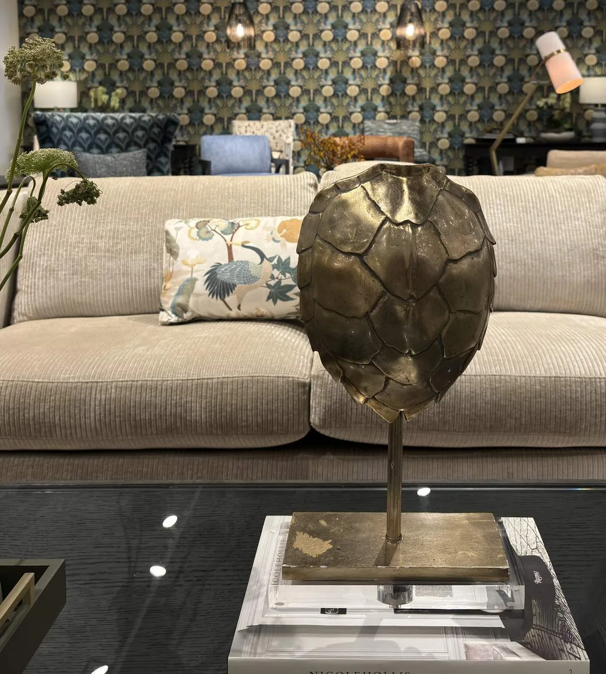 A living room with a beige corduroy sofa, decorative pillow, brass art piece on a glass coffee table, patterned wallpaper, and various chairs in the background