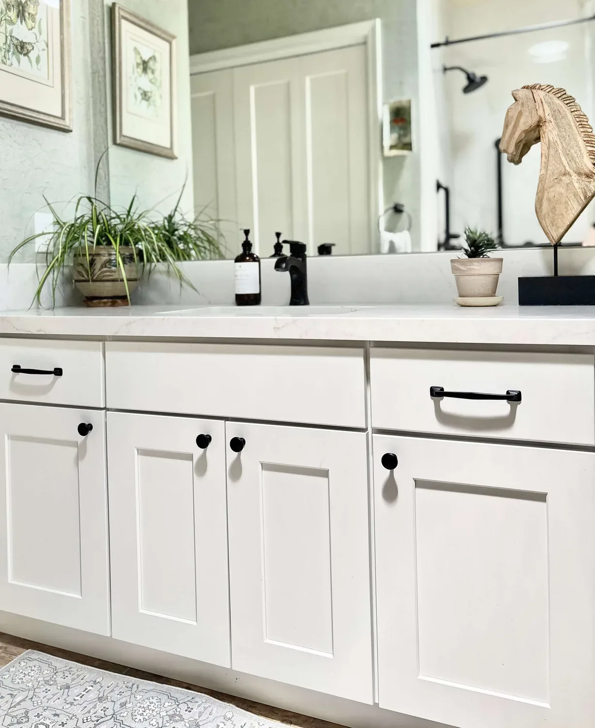 A white bathroom vanity with black hardware, a marble countertop, a black faucet, decorative plants, and a wooden horse