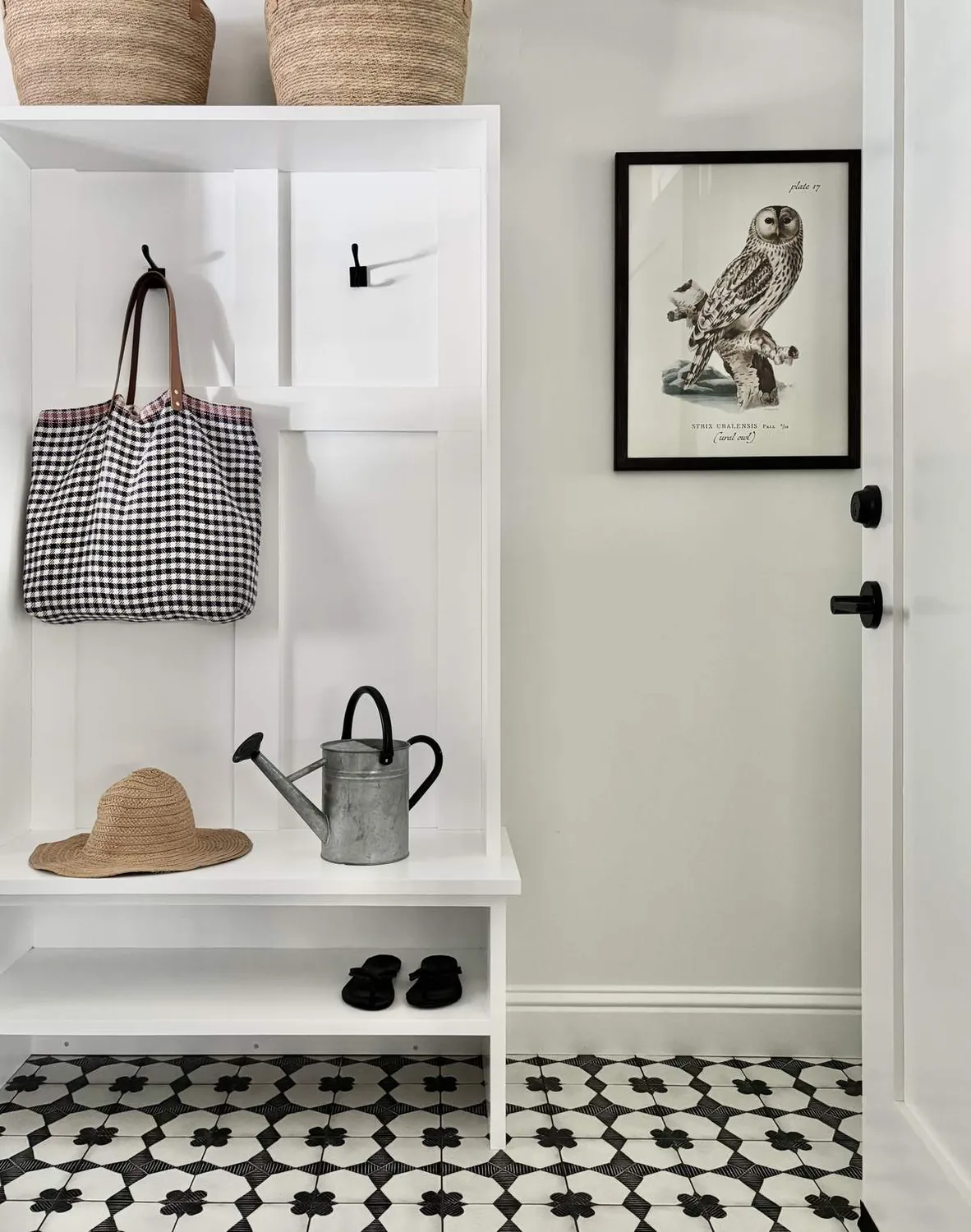 A minimalist entryway with a white bench, black and white patterned floor tiles, a framed owl print, and accessories