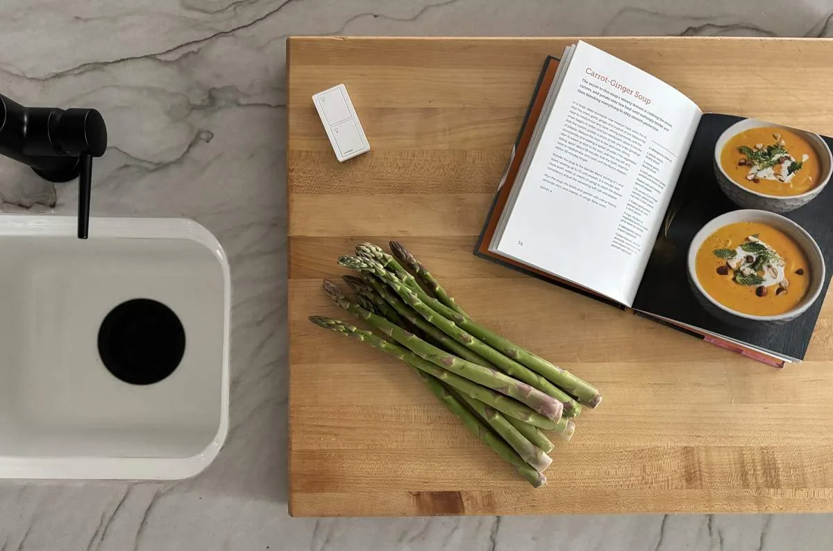 A kitchen countertop with a bundle of asparagus, an open cookbook showing a carrot-ginger soup recipe, and a white sink with a black faucet