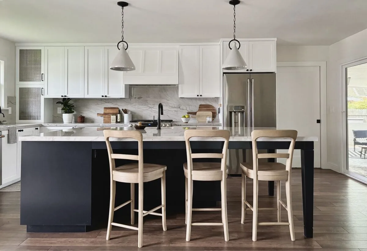 A modern kitchen with white upper cabinets, a black island with a white marble countertop, three wooden bar stools