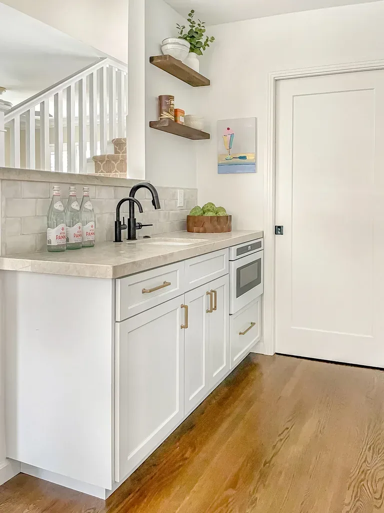 A modern kitchen area with white cabinetry, black faucet, wooden floating shelves, and a small built-in microwave