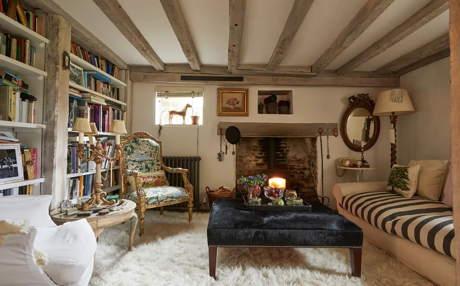 A cozy cottage living room with exposed ceiling beams, a brick fireplace, floor-to-ceiling bookshelves, a gilded armchair, and a tufted velvet ottoman