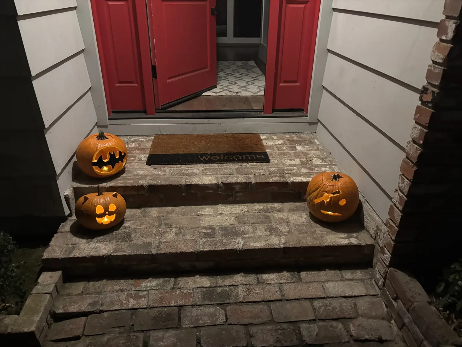 A front porch with red double doors, a welcome mat, and three carved, lit pumpkins on brick steps