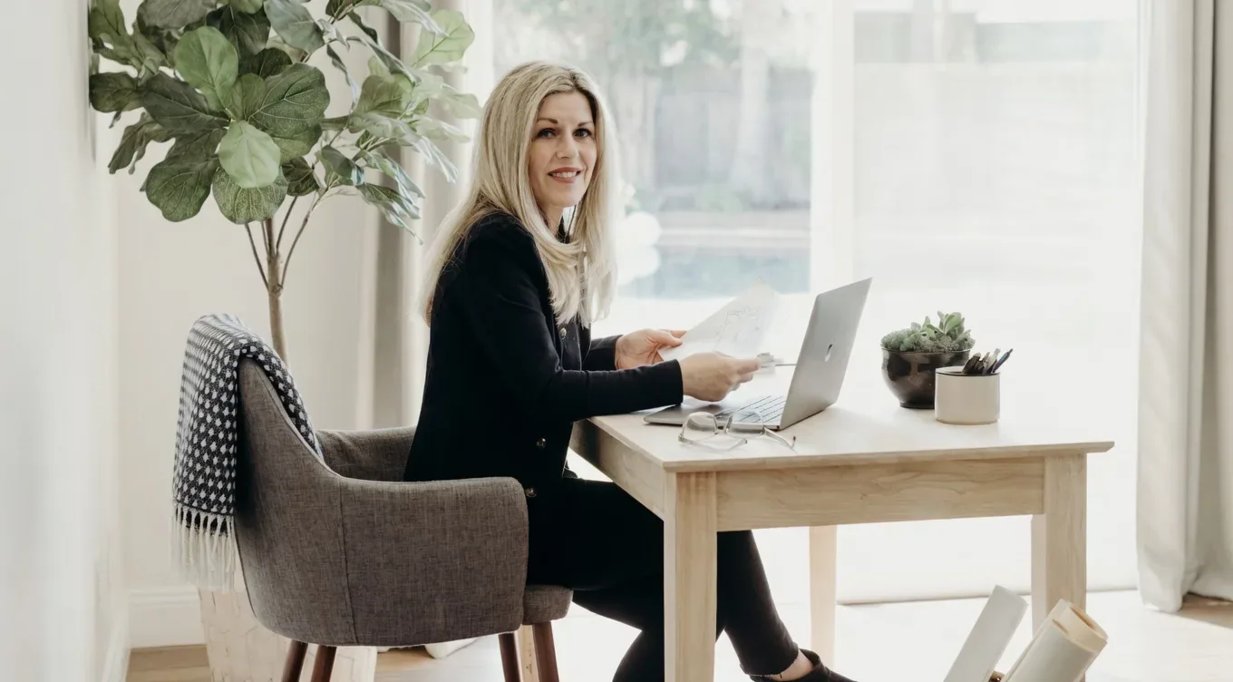 A woman sitting at a wooden desk in a bright, minimalist home office with a laptop, papers, and a plant