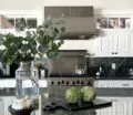 A modern kitchen with white cabinetry, a large island with a marble countertop, stainless steel appliances, and decorative plants