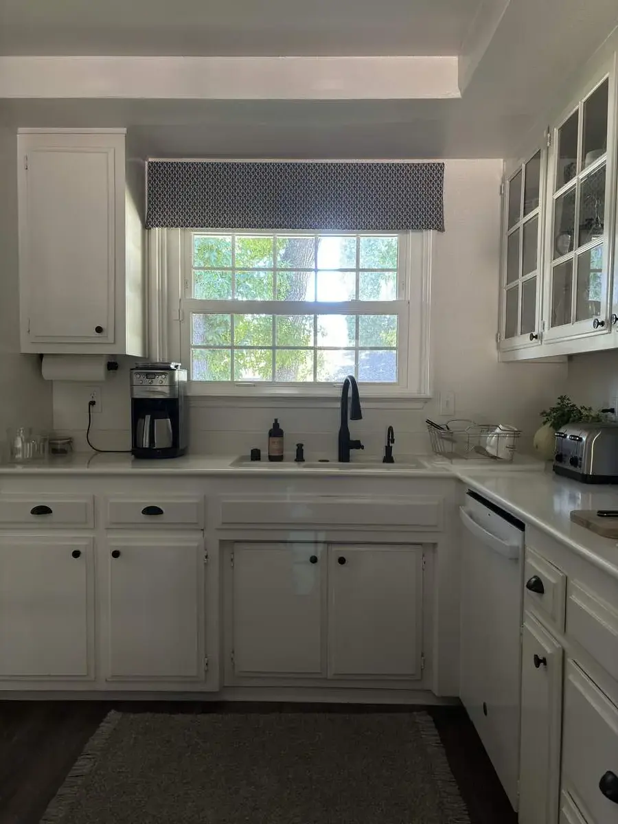 A bright kitchen with white cabinetry, a window with a patterned valance, a black faucet, and a coffee maker on the counter