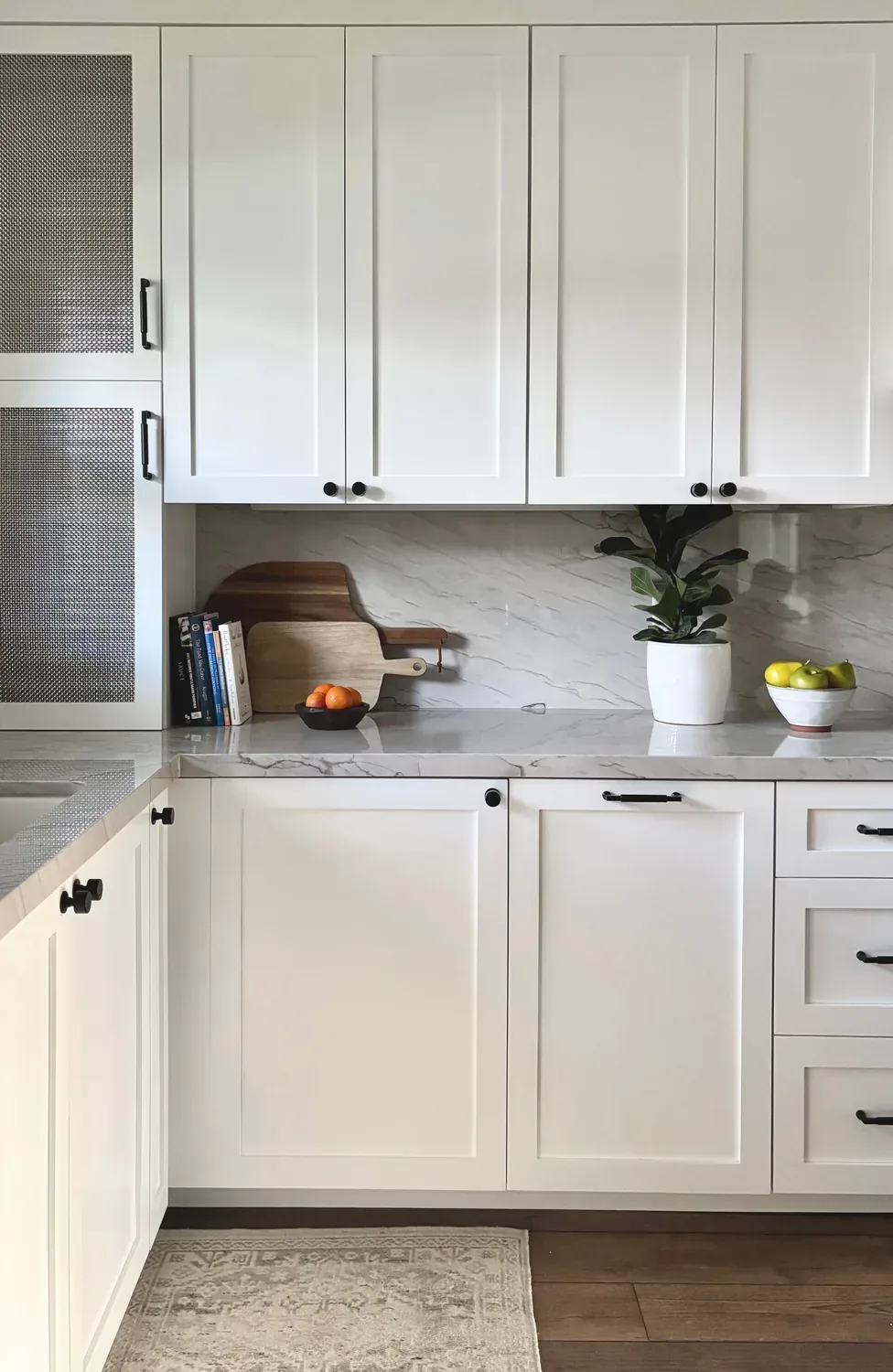 A modern kitchen with white cabinetry, marble countertops, a potted plant, cutting boards, and bowls of fruit