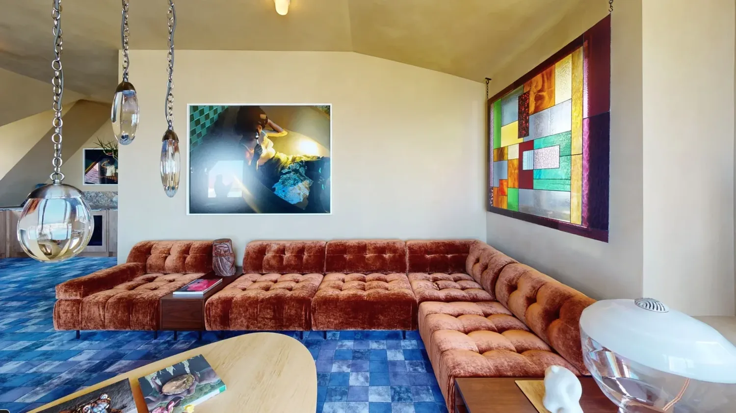 A living room with a large rust-colored tufted sectional sofa, blue checkered carpet, modern pendant lights, and colorful stained glass window