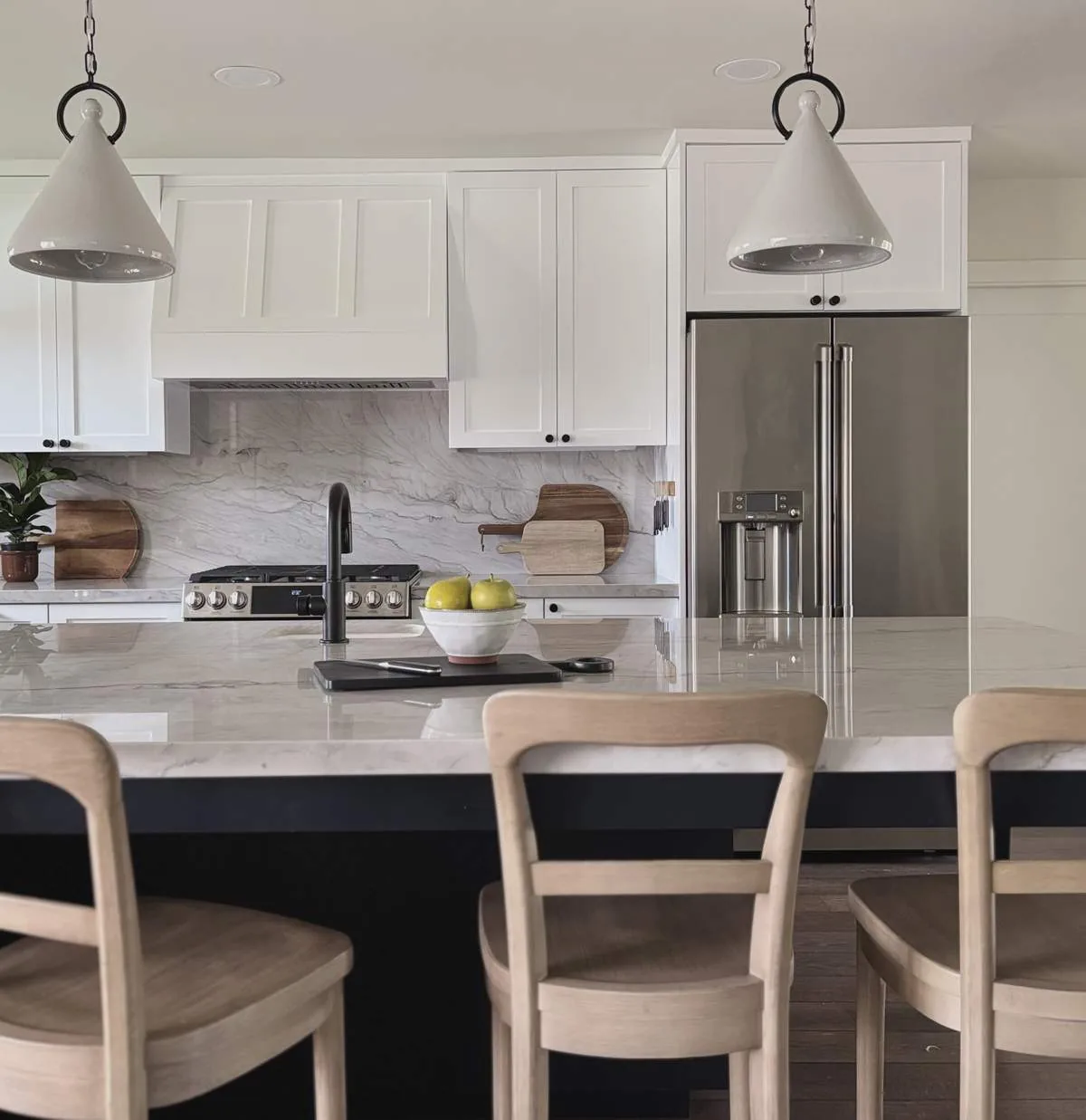 A modern kitchen with white cabinetry, marble countertops, stainless steel refrigerator, and wooden chairs at a kitchen island