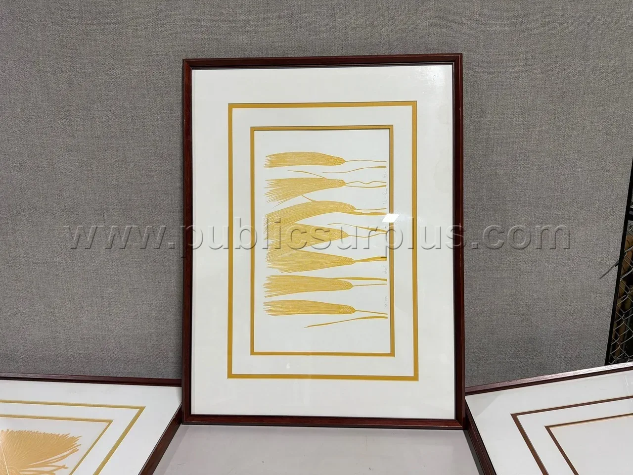 A framed abstract art print with yellow brushstroke designs on a white background, leaning against a gray fabric wall
