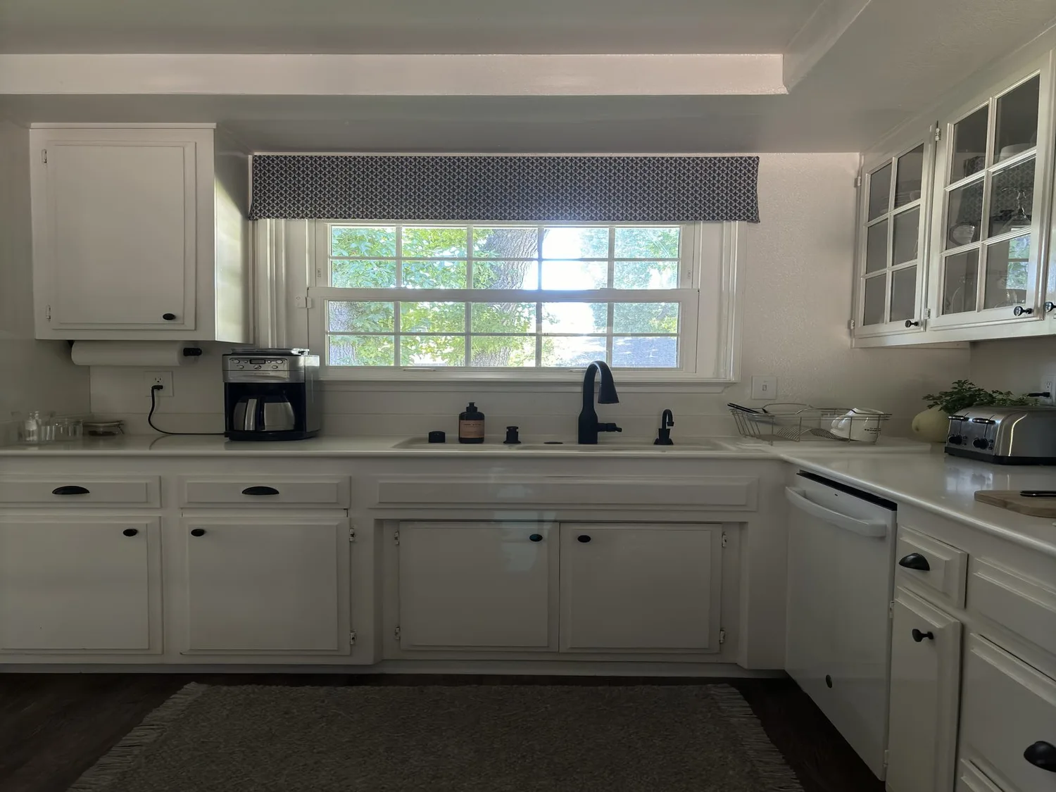 A bright remodeled kitchen with white cabinetry, a patterned window valance, a black faucet, and glass-front upper cabinets